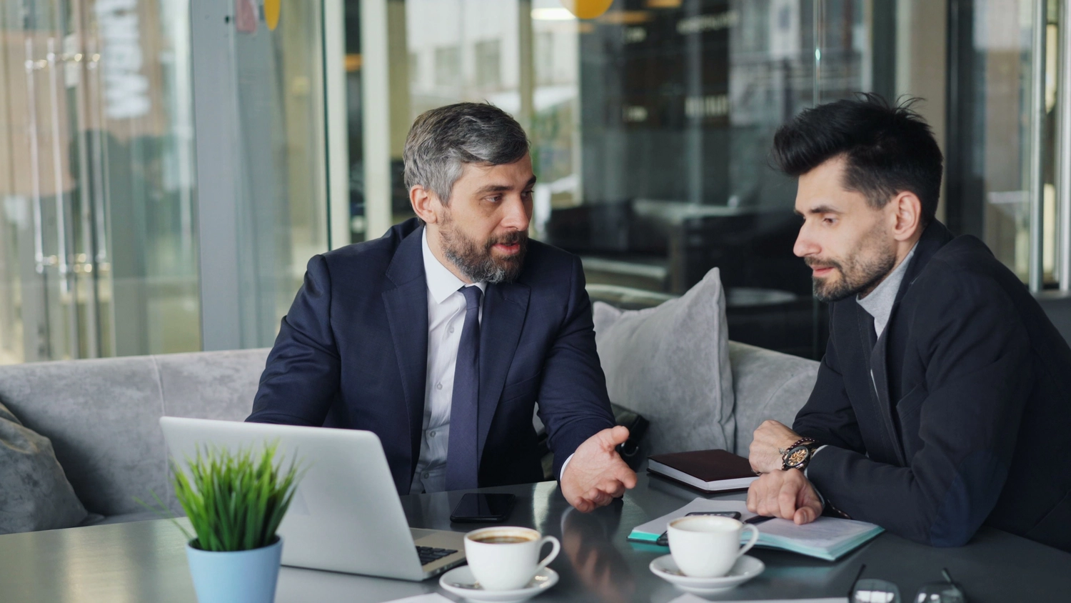 Simona Manoiu – Coaching und Psychotherapie – Dieses Bild zeigt zwei Männer in Businesskleidung, die sich an einem modernen Café-Tisch unterhalten.